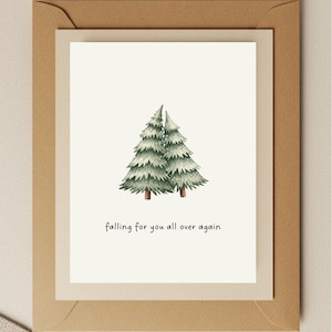 May include: A greeting card featuring two watercolor evergreen trees. The card has the phrase "falling for you all over again" in a handwritten font. The card is inside a cream-colored envelope.