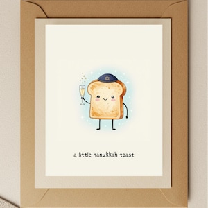 May include: A greeting card featuring a cartoon slice of toast wearing a blue yarmulke and holding a champagne flute. The text "a little hanukkah toast" is printed below the illustration. The card is set against a light blue background.