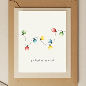 May include: Greeting card with a watercolor illustration of colorful string lights. The lights are red, green, blue, and yellow. The text "you light up my world" is written in black script below the lights. The card is inside a cream-colored envelope.