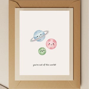 May include: Greeting card featuring a watercolor illustration of three smiling planets: a blue planet with rings, a pink planet, and a green planet. The text "you're out of this world!" is written below the planets. The card is inside a cream-colored envelope.