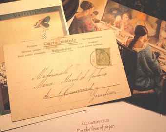 Op de afbeelding: Een verzameling ansichtkaarten en papier in vintage-stijl, waaronder een crèmekleurige ansichtkaart met cursief handschrift, een Vogue-tijdschriftomslag en een illustratie van een persoon in een blauw kledingstuk, met de tekst ALL CARDS CLUB.