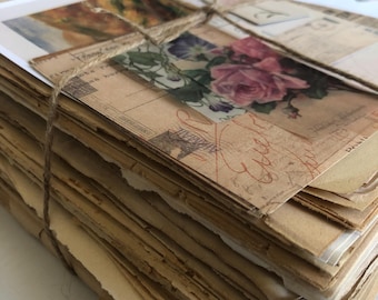 Op de afbeelding: Een stapel verouderd papier, vastgebonden met touw, met vintage ansichtkaarten en ephemera. De ansichtkaarten tonen bloemen- en landschapsbeelden, met vervaagde tekst en een sepia-tint. De bundel suggereert een verzameling antieke brieven of briefpapier.