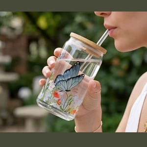 Könnte beinhalten: Ein klares Glas mit Bambusdeckel und Strohhalm, verziert mit einem blauen Schmetterling und floralem Muster. Das Glas ist mit Flüssigkeit gefüllt. Das Design umfasst rote und weiße Blumen.