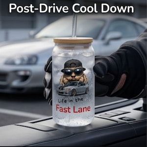 Könnte beinhalten: Ein klares Glas mit Bambusdeckel und Strohhalm, gefüllt mit Eis und Wasser. Das Glas zeigt eine Grafik einer Katze mit Sonnenbrille und Baseballkappe, darunter ein Auto. Der Text lautet "Life in the Fast Lane" und "Post-Drive Cool Down".