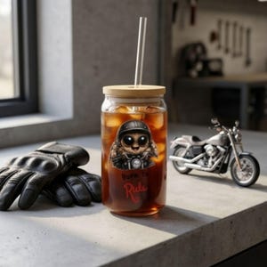 Könnte beinhalten: Ein klares Glas mit Bambusdeckel und Strohhalm, gefüllt mit dunkler Flüssigkeit und Eis. Das Glas zeigt eine Cartoon-Biker-Katze und den Text "Born To Ride". Schwarze Lederhandschuhe und ein Spielzeugmotorrad sind ebenfalls zu sehen.