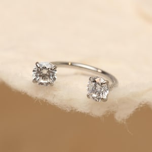 Peut inclure: Une bague argentée ornée de deux pierres précieuses claires, taillées en carré, placées aux extrémités opposées. La bague incurvée repose sur une surface douce et blanc cassé, créant une esthétique minimaliste et élégante. Les pierres scintillent.