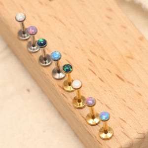 Pode incluir: Uma variedade de piercings labiais com pedras iridescentes tipo opala. Os piercings são exibidos em um bloco de madeira. Alguns piercings têm pinos prateados, enquanto outros são dourados. As pedras de opala são de várias cores, incluindo branco, azul e roxo.