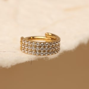 Puede incluir: Un pendiente de oreja dorado con tres filas de piedras transparentes y brillantes. La joya se encuentra sobre un fondo suave y blanquecino, resaltando su diseño. Este pendiente de oreja es un accesorio elegante.