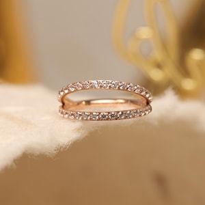 May include: A rose gold ring with two bands, each set with small, clear gemstones. The ring is displayed on a soft, off-white surface, with a blurred gold background.