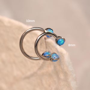 Puede incluir: Dos pendientes plateados en forma de herradura con piedras azules similares a ópalos. Uno mide 10 mm de diámetro, el otro 8 mm. Diseñados para piercings.