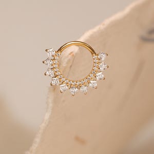 May include: A gold septum ring with a sunburst design. The ring features square and round clear gemstones. The jewellery is set against a neutral background.