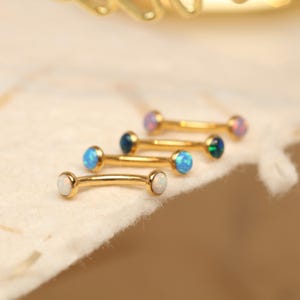 May include: Gold-colored curved barbell earrings with opal and gemstone accents. The earrings feature a curved bar with a small gemstone at each end. The gemstones are in various colors, including white, blue, and purple.