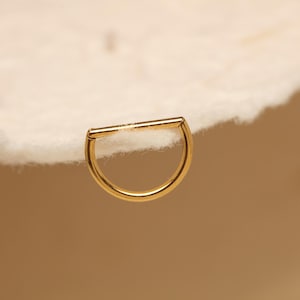 Könnte beinhalten: Ein goldfarbener Septumring mit Scharnierdesign. Der Ring hat eine halbkreisförmige Form mit einem geraden Steg oben. Der Schmuck wird vor einem weichen, cremefarbenen Hintergrund gezeigt.