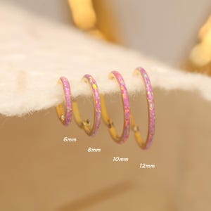 Könnte beinhalten: Vier goldene Creolen mit rosa Opal-Inlays, in Größen von 6 mm bis 12 mm. Die Ohrringe werden auf einer weichen, weißen Oberfläche präsentiert. Die Größen sind auf dem Bild beschriftet.