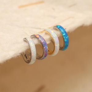May include: Five hoop earrings with iridescent opal-like inlays. The earrings are silver, gold, and blue. The opal inlays have a range of colours, including white, purple, and blue. The earrings are arranged in a row.