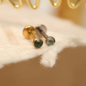 Puede incluir: Dos pendientes pequeños de botón. Uno es dorado, el otro plateado. Cada uno tiene una pequeña piedra verde redonda. Los pendientes están sobre una superficie blanca texturizada. Los pendientes son probablemente para piercings en la oreja o el labio.
