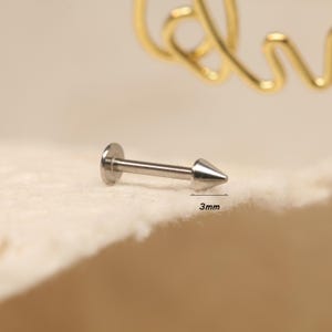 May include: A silver-colored, spiked stud earring. The earring has a flat, round backing and a conical, pointed end. The image shows the earring on a light-colored, textured surface. The text "3mm" is next to the earring.