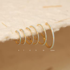 Può includere: Una collezione di orecchini a cerchio dorati con intarsi di opale, presentati in dimensioni crescenti. Gli orecchini variano da 6 mm a 12 mm di diametro, offrendo una varietà di opzioni. Gli intarsi di opale aggiungono un tocco di iridescenza ai cerchi dorati.