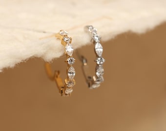 May include: Two hoop earrings, one gold-toned and one silver-toned, each featuring a line of clear, sparkling gemstones. The gemstones alternate between round and marquise shapes, creating a delicate and elegant design.