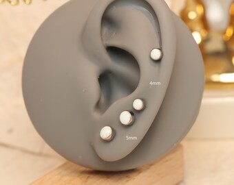 Puede incluir: Exhibición de pendientes de botón en un modelo de oreja gris. Los pendientes son plateados con centros circulares blancos. Los tamaños están etiquetados como 4 mm y 5 mm. El modelo está colocado sobre un bloque de madera.