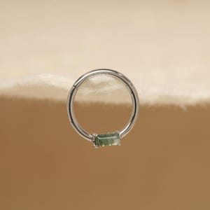 May include: A silver-colored hinged ring with a rectangular, light green gemstone. The ring is set against a neutral background, highlighting the jewelry's design and color.