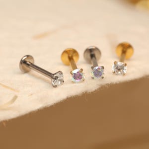 May include: Four small, stud earrings with a flat back and a small, round, sparkling gem. Two earrings have a gold post, one has a silver post, and one has a silver post. The gems are clear and iridescent.