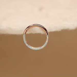 Könnte beinhalten: Roségoldener Klappcreolen-Ohrring mit weißem Opal-Inlay. Der Ohrring ist kreisförmig und hat ein nahtloses Design. Das Opal-Inlay hat eine subtile, irisierende Qualität. Der Ohrring wird vor neutralem Hintergrund gezeigt.