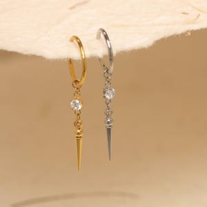 May include: Two hoop earrings, one gold-toned and one silver-toned, each with a dangling design. The gold earring features a clear gemstone and a spike pendant. The silver earring has a clear gemstone and a spike pendant. The earrings are displayed against a neutral background.