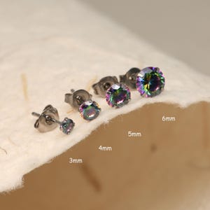May include: Five pairs of iridescent rainbow stud earrings in different sizes: 3mm, 4mm, 5mm, and 6mm. The earrings are made of stainless steel and have a black finish.