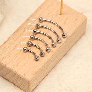 Può includere: Una collezione di piercing curvi per sopracciglia, di colore argento, esposti su un blocco di legno chiaro. Le misure, da 6 mm a 16 mm, sono indicate. Ogni piercing è di tipo barbell, con una sfera a ciascuna estremità.