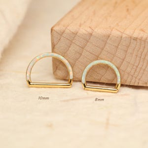 May include: Two gold-colored, half-circle shaped earrings with a white opal inlay. One earring is labeled 10mm, the other 8mm. The earrings are displayed on a light-colored wooden surface.