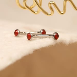 May include: Two silver-colored barbell earrings with red gemstone ends. The earrings are displayed on a white, textured surface. A gold-colored, abstract metal design is visible in the background.