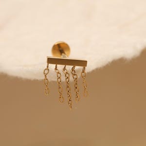May include: A gold-colored earring with a post and a rectangular bar design. Five delicate chains hang from the bar, creating a dangling effect. The earring is set against a soft, off-white background.