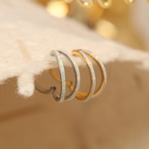 Pode incluir: Dois pares de brincos de argola. Um par é prateado com uma incrustação de opala branca, e o outro é dourado com uma incrustação de opala branca. Os brincos são exibidos em uma superfície esbranquiçada.
