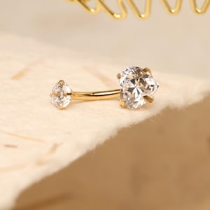 Pode incluir: Um brinco de barra curva dourado com duas pedras de zircônia cúbica transparentes e brilhantes. Uma pedra tem formato de gota e a outra de coração. O brinco está sobre um fundo neutro.