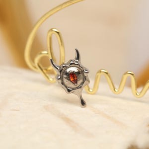 May include: A silver-toned pendant with a red gemstone center, featuring a unique, stylized design with curved horns and a teardrop shape. The pendant is set against a gold-colored, looping wire.