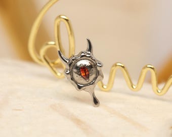 May include: A silver-toned pendant with a red gemstone center, featuring a unique, stylized design with curved horns and a teardrop shape. The pendant is set against a gold-colored, looping wire.