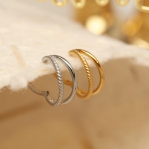 Könnte beinhalten: Nahaufnahme von zwei Doppel-Ohrringen. Ein Ohrring ist silberfarben mit einem gedrehten Seildesign am äußeren Reifen. Der andere Ohrring ist goldfarben mit einem ähnlichen gedrehten Seildesign. Die Ohrringe liegen auf einer hellen, strukturierten Oberfläche.