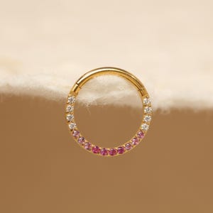 May include: A gold-colored hinged hoop earring. The earring is adorned with a gradient of gemstones, transitioning from clear to pink. The gemstones are evenly spaced around the hoop's circumference, creating a sparkling effect.