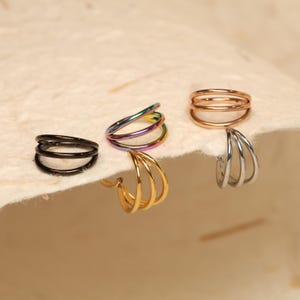 May include: A selection of hoop earrings in black, gold, silver, and iridescent finishes. Each earring has a triple-band design. The earrings are presented on a textured, off-white surface.