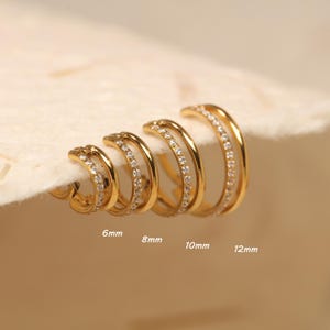 Puede incluir: Un conjunto de pendientes de aro dorados en varios tamaños, cada uno con un diseño de doble banda. Una banda está adornada con pequeñas gemas transparentes. Los tamaños son 6 mm, 8 mm, 10 mm y 12 mm.