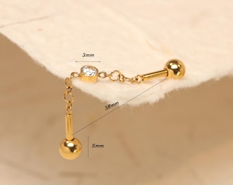 Op de afbeelding: Een goudkleurige oorbel met een bungelende ketting en een kleine, ronde, heldere edelsteen. De oorbel is 3,8 cm lang, met een bungelende bal van 5 mm. De edelsteen heeft een diameter van 3 mm.