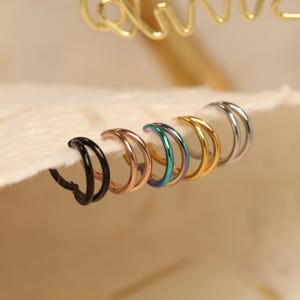 Könnte beinhalten: Eine Auswahl an Doppel-Creolen in verschiedenen Farben, darunter Schwarz, Roségold, schillernd, Gold und Silber. Die Ohrringe werden auf einer weichen, cremefarbenen Oberfläche präsentiert, mit einem verschwommenen goldenen Dekorationselement im Hintergrund.