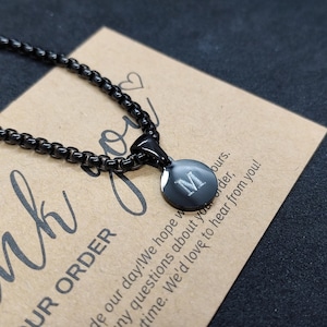 May include: A black necklace with a circular pendant featuring the letter "M". The pendant and chain are dark in color. The necklace rests on a brown card with the words "UR ORDER" and other text.