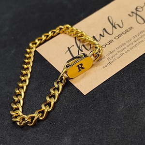 May include: A gold-coloured chain bracelet with a rectangular clasp bearing the letter "R". The bracelet, made of interlocking links, lies on a brown card with the words "thank you" and "OUR ORDER".