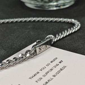 May include: A silver chain necklace with a clasp that has the letters "S" and "A" engraved on it, along with a small heart symbol. The necklace is lying on a white card with the text "Thank you so much for supporting my small business."