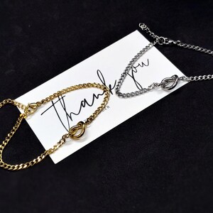May include: Two chain bracelets, one gold-toned and one silver-toned, are displayed on a white card with the word "Thank You" written on it. The bracelets feature a knot design. The background is black.