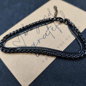 Peut inclure: Un bracelet chaîne noir avec un motif texturé repose sur une carte marron. La carte porte les mots "be kind & grateful". Le bracelet est en métal foncé et possède une fermeture à fermoir.