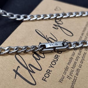 May include: A silver curb chain necklace with a silver clasp that says "TCB" on it. The necklace is lying on a brown card with the text "Thank you for your order" written on it.