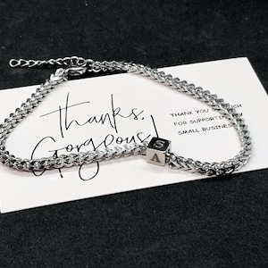 Puede incluir: Una pulsera de cadena plateada con un pequeño dije cuadrado con las letras "A" y "S". La pulsera está sobre una tarjeta blanca con las palabras "thanks gorgeous" y "THANK YOU SO MUCH FOR SUPPORTING SMALL BUSINESSES".
